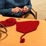 A person's hands with red yarn and a knitted red hat with a tassel